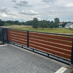 Usługi spawalnicze Jakub Śledź - Przesuwna brama z poziomych desek w kolorze brązowym, osadzona w czarnej metalowej ramie, na tle wiejskiego krajobrazu z polami i turbinami wiatrowymi.