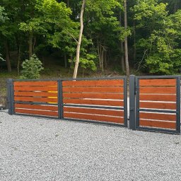 Usługi spawalnicze Jakub Śledź - Nowoczesna brama wjazdowa i furtka z poziomych desek w kolorze ciepłego drewna, osadzone w grafitowej, metalowej ramie, na tle zielonego lasu i szarego żwiru.