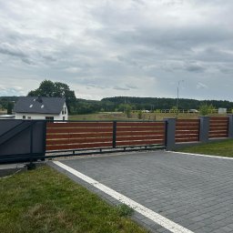 Usługi spawalnicze Jakub Śledź - Przesuwna brama wjazdowa z drewnianymi panelami i szarą metalową konstrukcją, widok na posesję z kostką brukową i dom w tle.