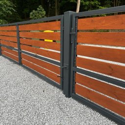 Usługi spawalnicze Jakub Śledź - Nowoczesna brama wjazdowa z szarymi metalowymi ramami i poziomymi deskami z drewna w kolorze ciepłego brązu, widok na żwirowe podłoże i zieleń w tle.