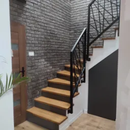 Drewniane schody z czarną metalową balustradą w nowoczesnym wnętrzu z ceglaną ścianą i drewnianą podłogą.