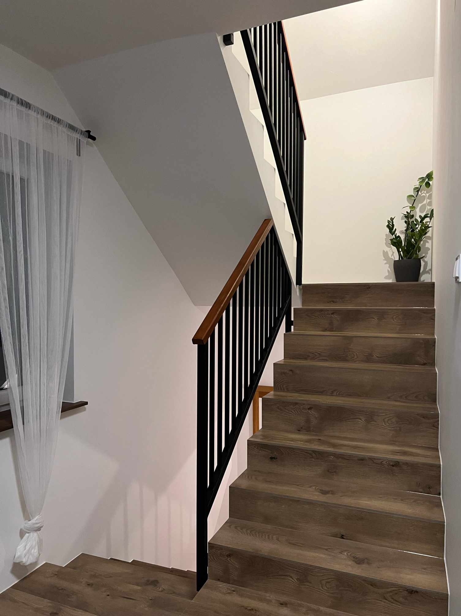 Schody wewnętrzne z drewnopodobnymi stopniami i czarną balustradą z drewnianym poręczą, oświetlone naturalnym światłem z okna z białą firanką.