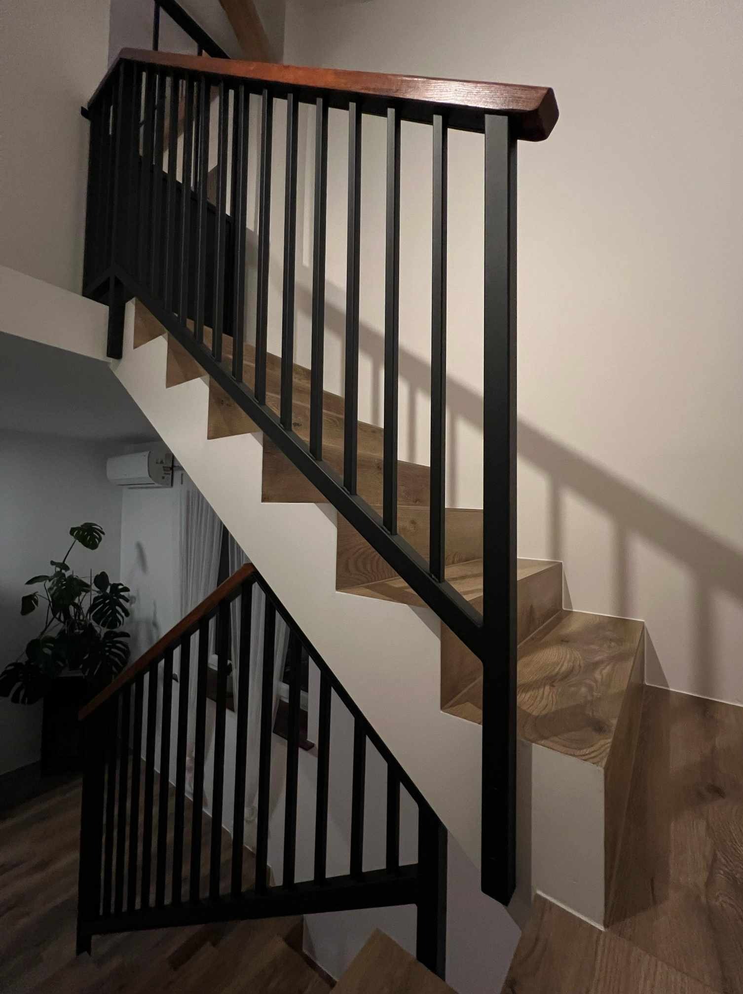 Wewnętrzne schody z drewnianymi stopniami i czarną metalową balustradą z drewnianym poręczem, widok z dołu, w tle biała ściana i roślina doniczkowa.