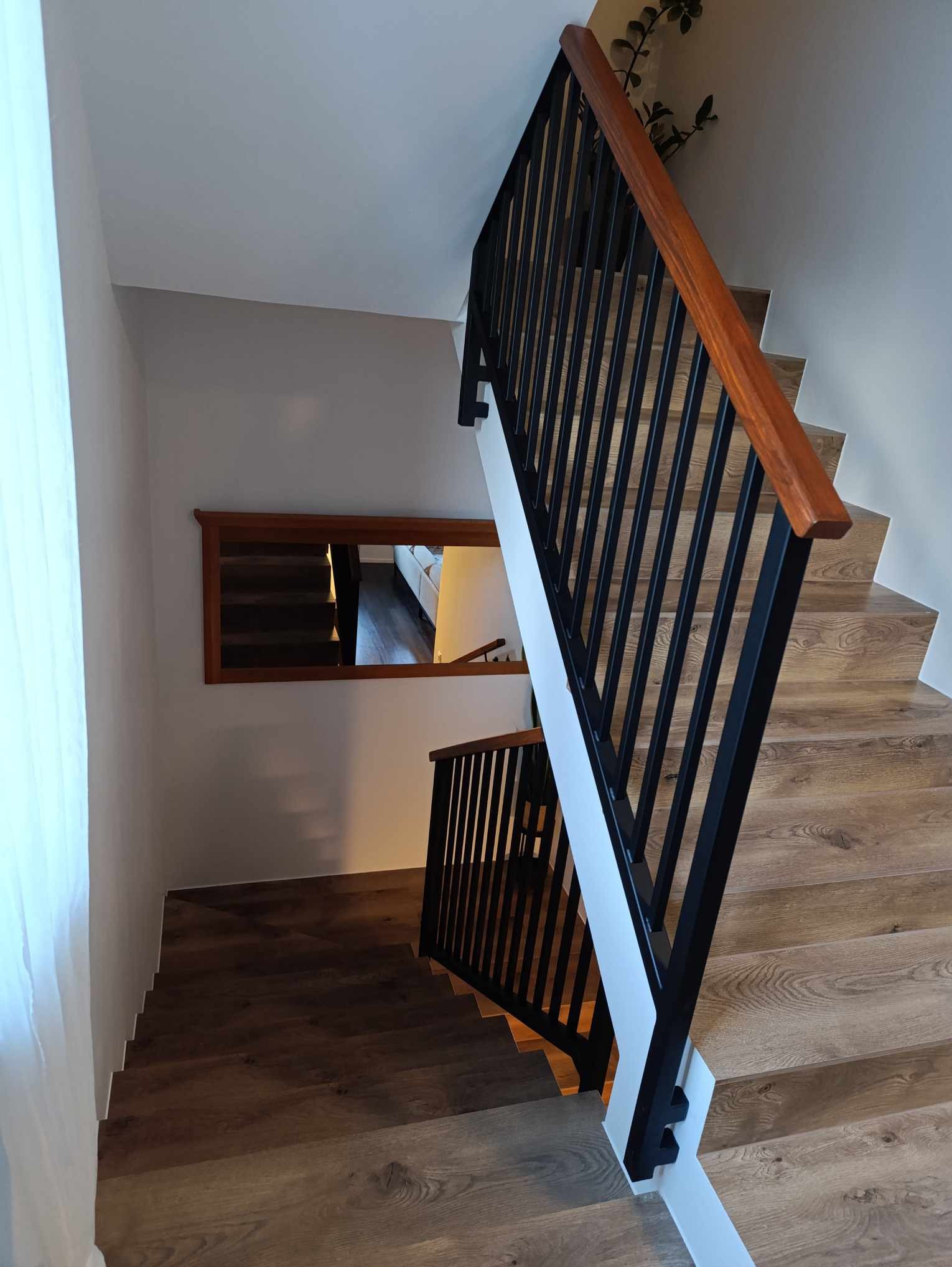 Wnętrze domu ze schodami drewnianymi z czarną metalową balustradą i drewnianą poręczą, widok z góry, na ścianie lustro odbijające dalszą część schodów.