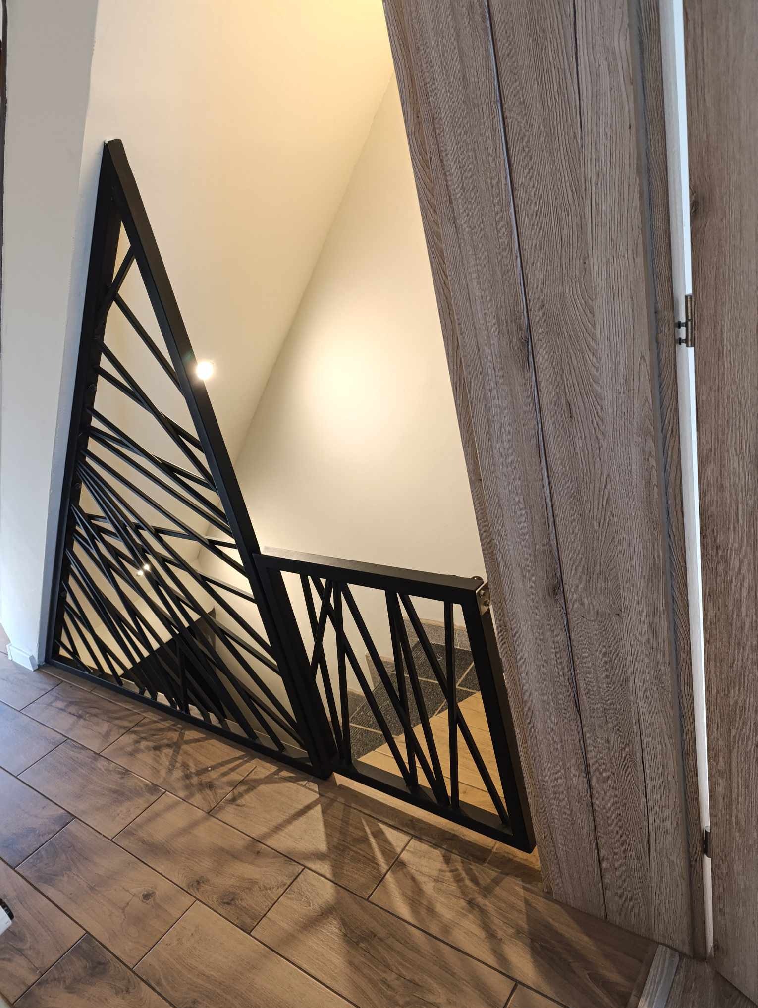 Nowoczesna, czarna balustrada schodowa z geometrycznym wzorem ukośnych prętów, widziana z dołu schodów, z fragmentem drewnianej podłogi i drzwi w tle.