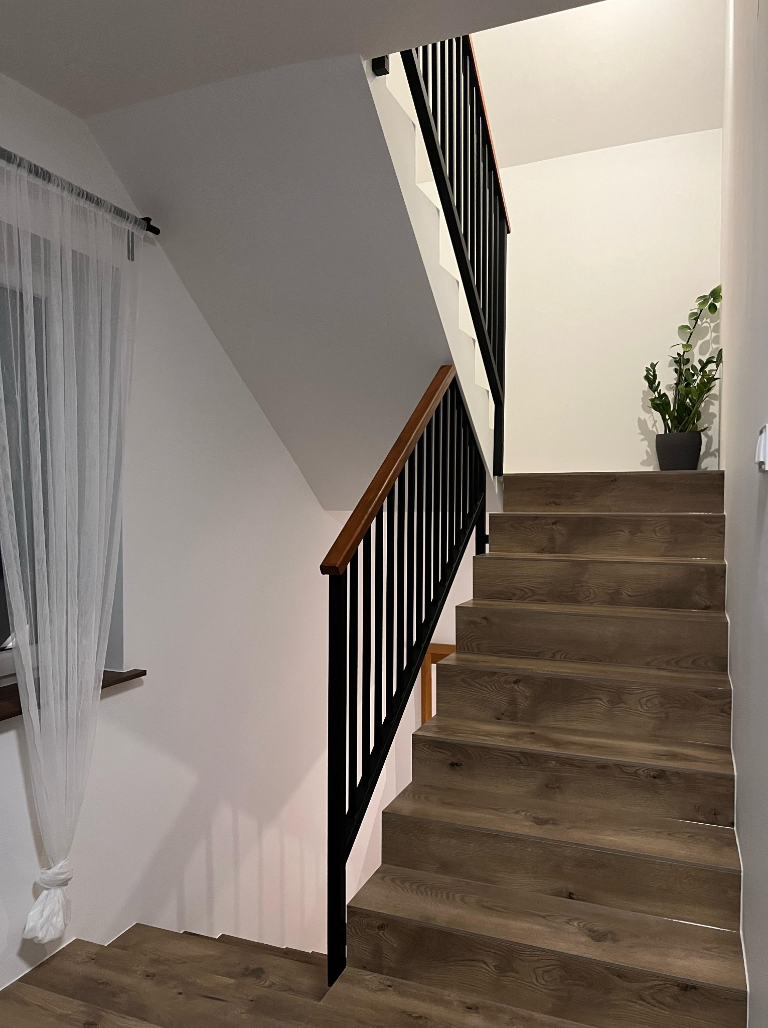 Wnętrze domu ze schodami wyłożonymi panelami imitującymi drewno, czarna balustrada z drewnianym poręczem, okno z białą firanką i doniczka z zieloną rośliną na górze schodów.