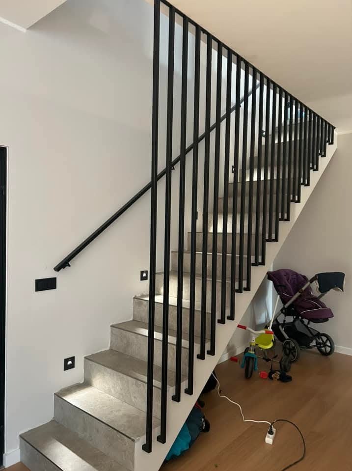 Schody wewnętrzne z szarymi stopniami i czarną metalową balustradą z pionowymi szczeblami w nowoczesnym domu, widoczne czarne włączniki światła i dziecięcy wózek w tle.