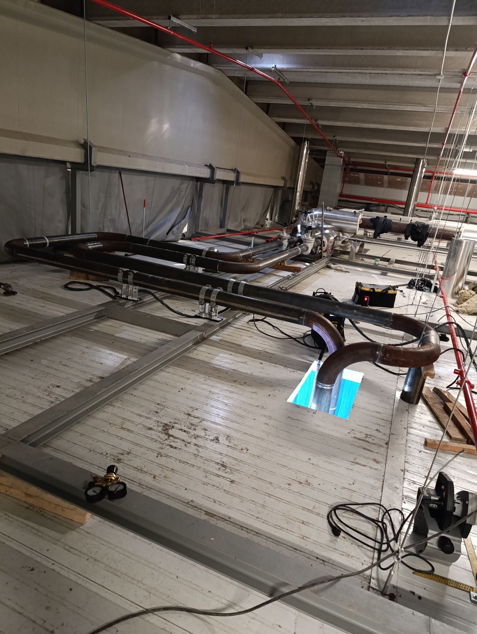 Spawanie rur metalowych w hali przemysłowej, widoczne spawane elementy, narzędzia i osprzęt spawalniczy, całość na tle konstrukcji stalowej i instalacji pod stropem.