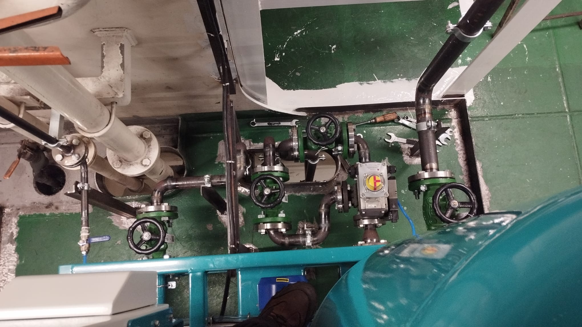 Instalacja przemysłowa z rurami i zaworami, widoczne narzędzia monterskie, fragment buta osoby fotografującej w dolnej części kadru, zielona podłoga i fragment białej ściany.