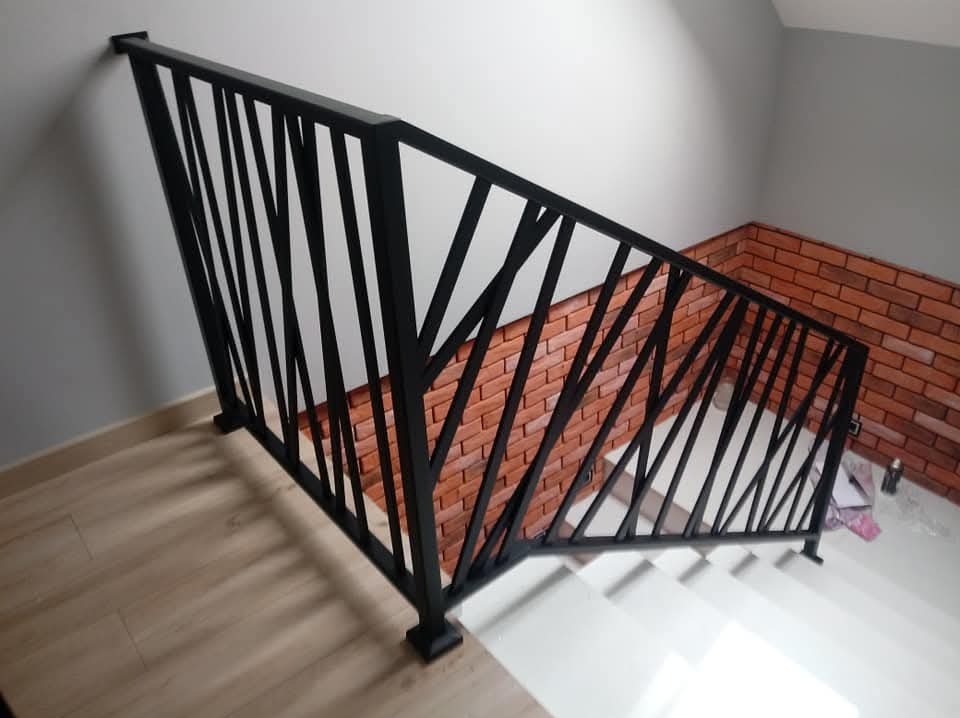 Czarna, metalowa balustrada schodowa z ukośnymi elementami, na tle ściany wyłożonej cegłą klinkierową, widok z góry.