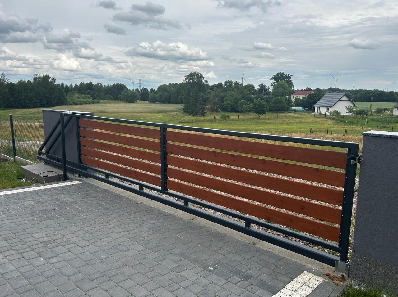 Przesuwna brama z poziomych desek w kolorze brązowym, osadzona w czarnej metalowej ramie, na tle wiejskiego krajobrazu z polami i turbinami wiatrowymi.
