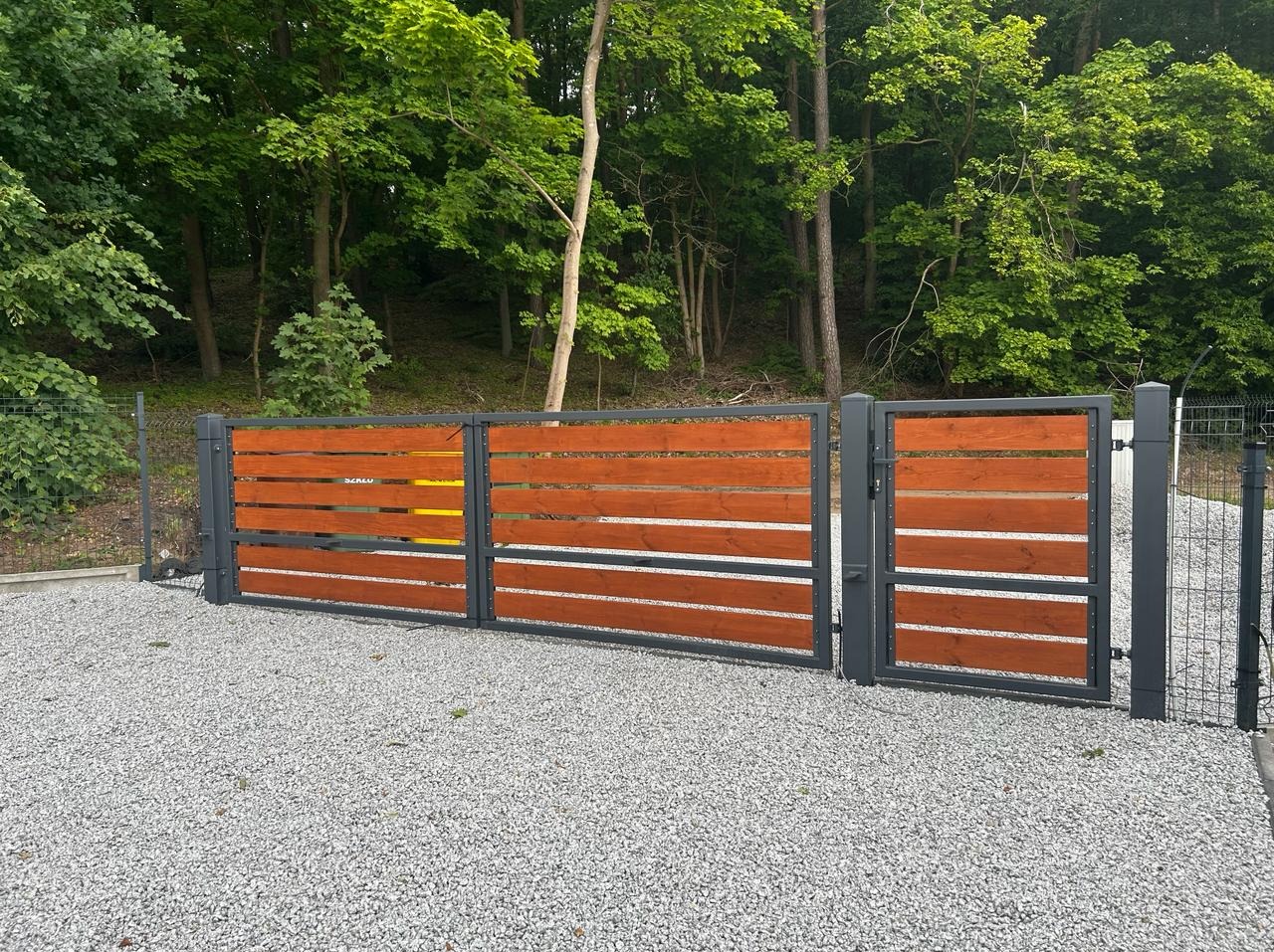 Nowoczesna brama wjazdowa i furtka z poziomych desek w kolorze ciepłego drewna, osadzone w grafitowej, metalowej ramie, na tle zielonego lasu i szarego żwiru.