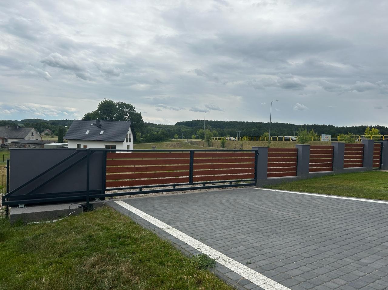 Przesuwna brama wjazdowa z drewnianymi panelami i szarą metalową konstrukcją, widok na posesję z kostką brukową i dom w tle.