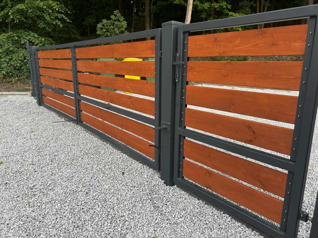 Nowoczesna brama wjazdowa z szarymi metalowymi ramami i poziomymi deskami z drewna w kolorze ciepłego brązu, widok na żwirowe podłoże i zieleń w tle.