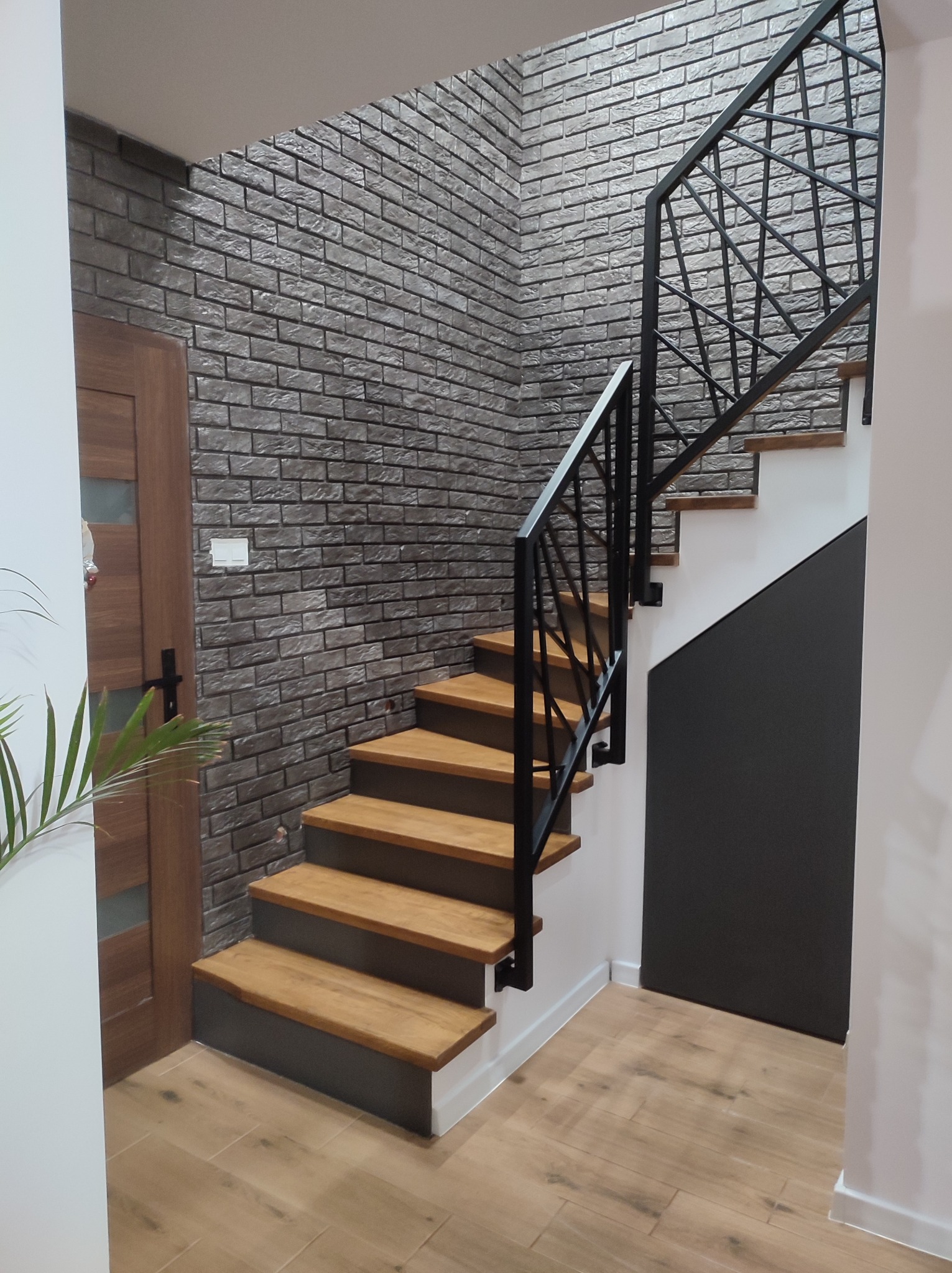 Drewniane schody z czarną metalową balustradą w nowoczesnym wnętrzu z ceglaną ścianą i drewnianą podłogą.