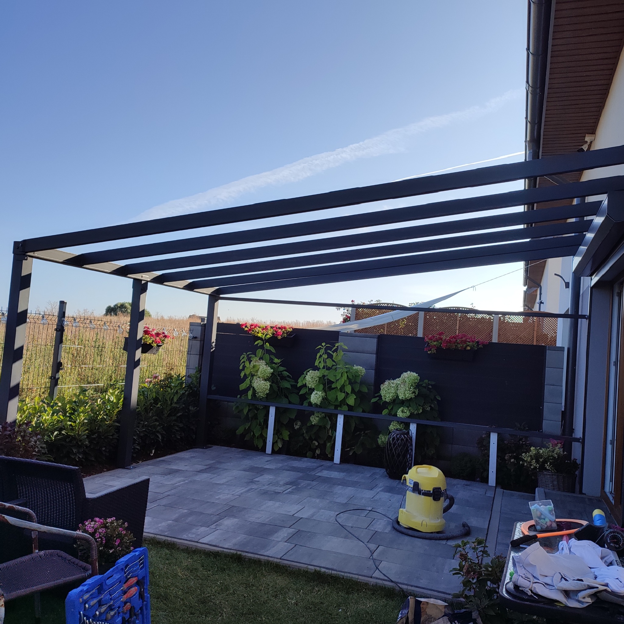 Nowoczesna pergola z metalową konstrukcją i poziomymi belkami, z kwitnącymi roślinami w tle i ułożonym brukiem na podłożu, sfotografowana w słoneczny dzień.