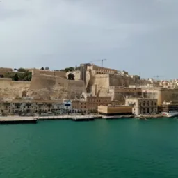 Panoramiczny widok na port w Valletcie, Malta, z widocznymi zabytkowymi budynkami, murami obronnymi i turkusową wodą, z dźwigiem budowlanym w tle.