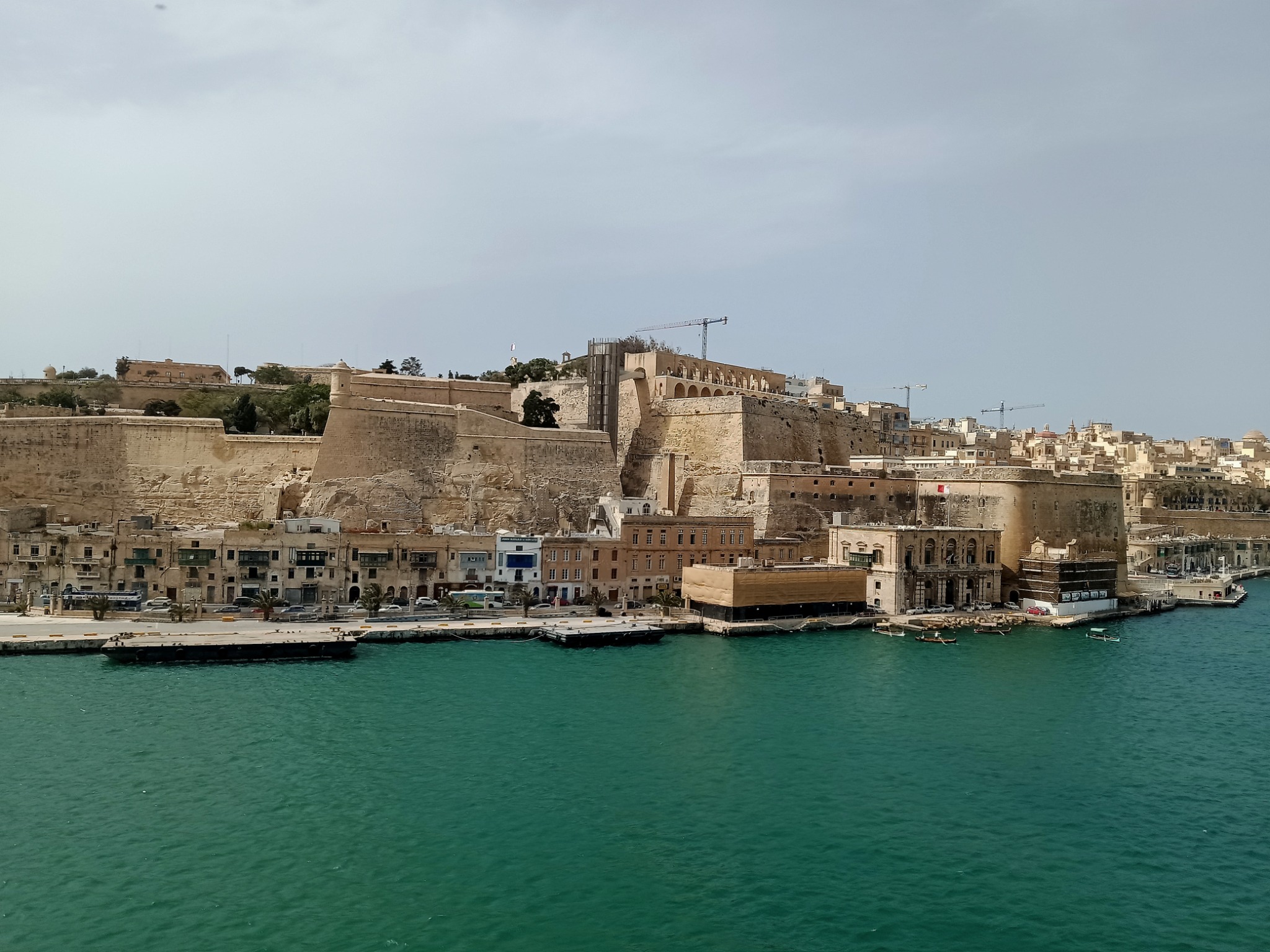 Panoramiczny widok na port w Valletcie, Malta, z widocznymi zabytkowymi budynkami, murami obronnymi i turkusową wodą, z dźwigiem budowlanym w tle.