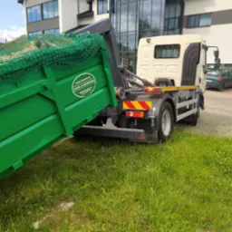 Zielony kontener transportowany przez ciężarówkę z widocznym logo firmy transportowej na tle nowoczesnego budynku.