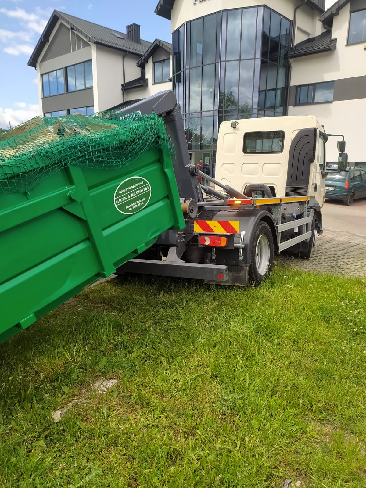 Zielony kontener transportowany przez ciężarówkę z widocznym logo firmy transportowej na tle nowoczesnego budynku.