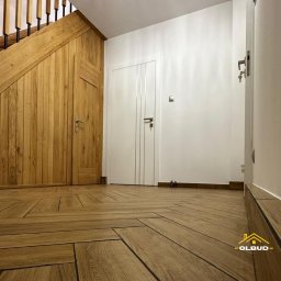 OLBUD Bartosz Gliński - Wykończony korytarz z drewnianą podłogą ułożoną w jodełkę, zabudową schodów z drewna i białymi drzwiami. Widoczny fragment schodów i listwy przypodłogowe.
