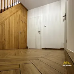 Wykończony korytarz z drewnianą podłogą ułożoną w jodełkę, zabudową schodów z drewna i białymi drzwiami. Widoczny fragment schodów i listwy przypodłogowe.