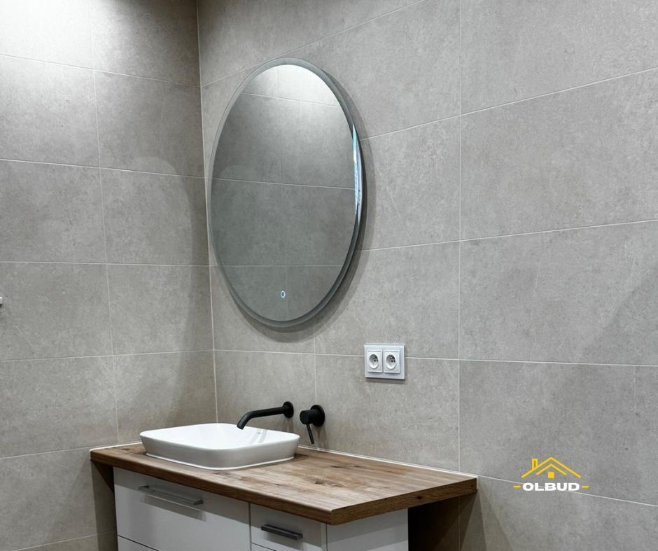 Wykończona łazienka z owalnym lustrem, umywalką nablatową i drewnianym blatem. Szare płytki na ścianach, czarna bateria i białe szafki pod umywalką. Nowoczesny design.