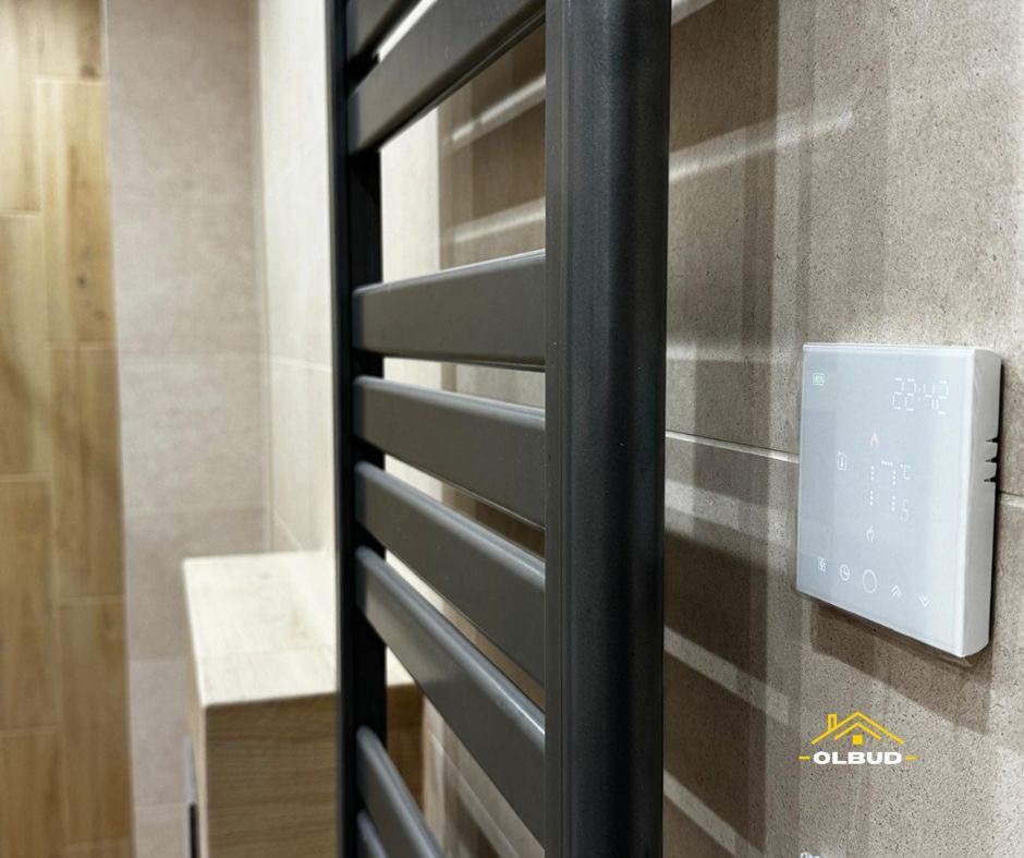 Nowoczesny, czarny grzejnik łazienkowy z elektronicznym sterownikiem na tle beżowych płytek. Minimalistyczny design, precyzyjny montaż.