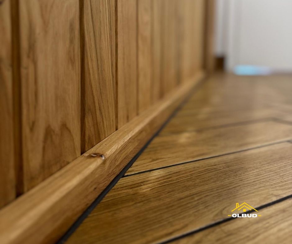 Drewniana boazeria i parkiet w jodełkę z czarnymi akcentami. Detal wykończenia wnętrza, ciepłe oświetlenie.