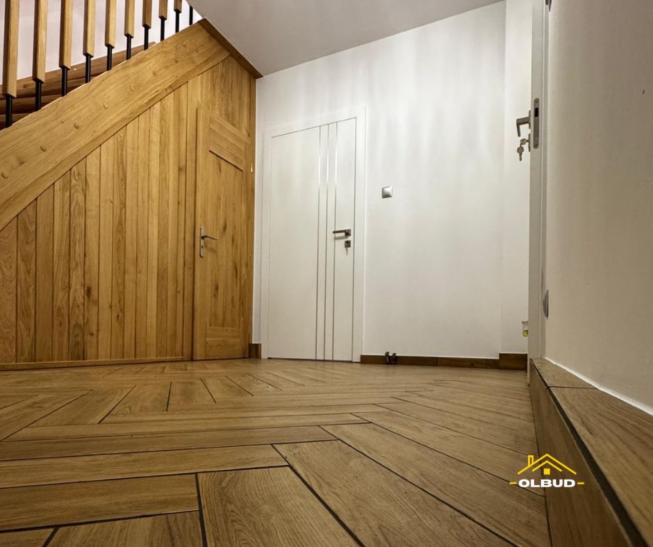 Wykończony korytarz z drewnianą podłogą ułożoną w jodełkę, zabudową schodów z drewna i białymi drzwiami. Widoczny fragment schodów i listwy przypodłogowe.