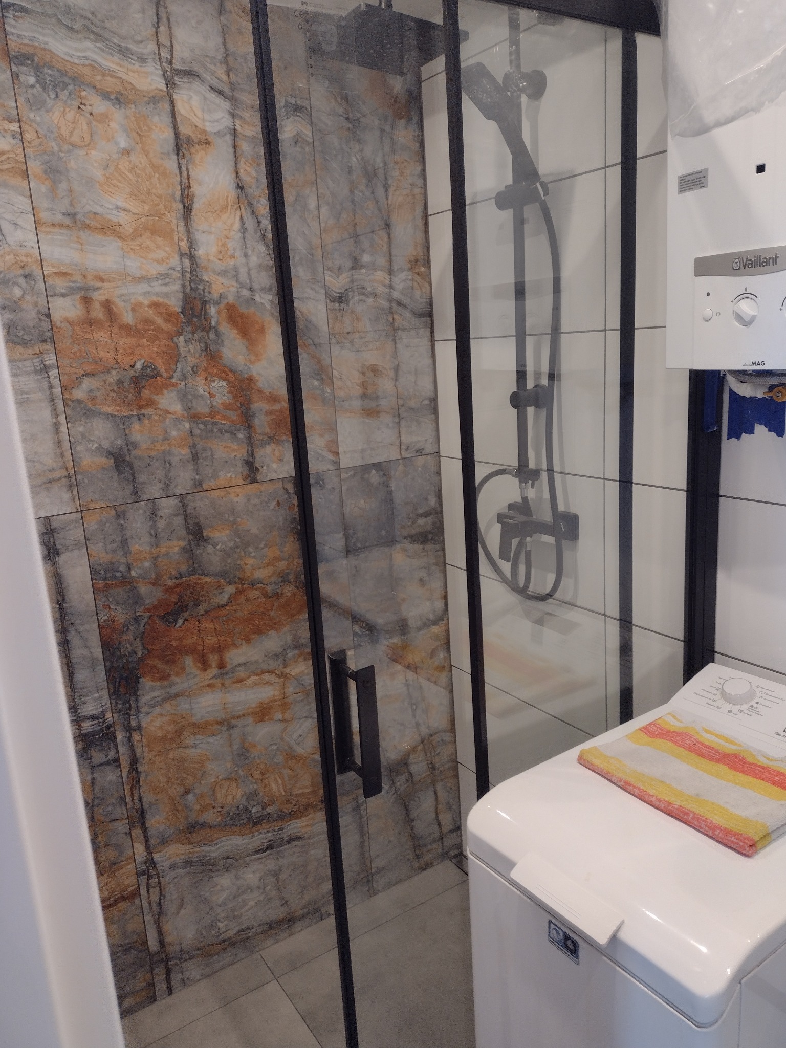 Kabina prysznicowa z czarnymi profilami i drzwiami ze szkła, obok pralka z ręcznikiem w paski, na ścianie płytki imitujące marmur oraz piec gazowy Vaillant.