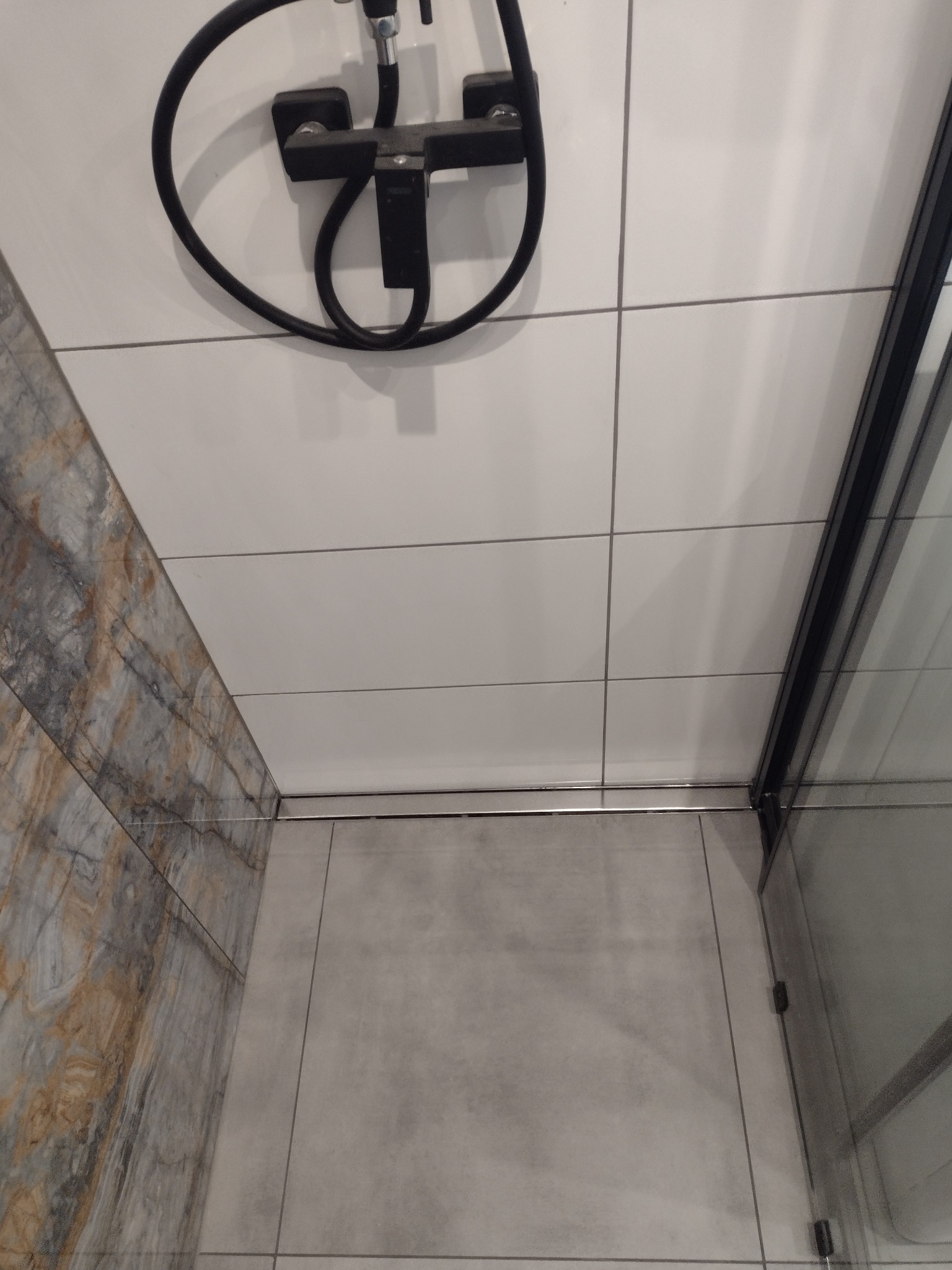 Nowoczesna kabina prysznicowa z czarną armaturą i odpływem liniowym, wykończona białymi i marmurkowymi płytkami.