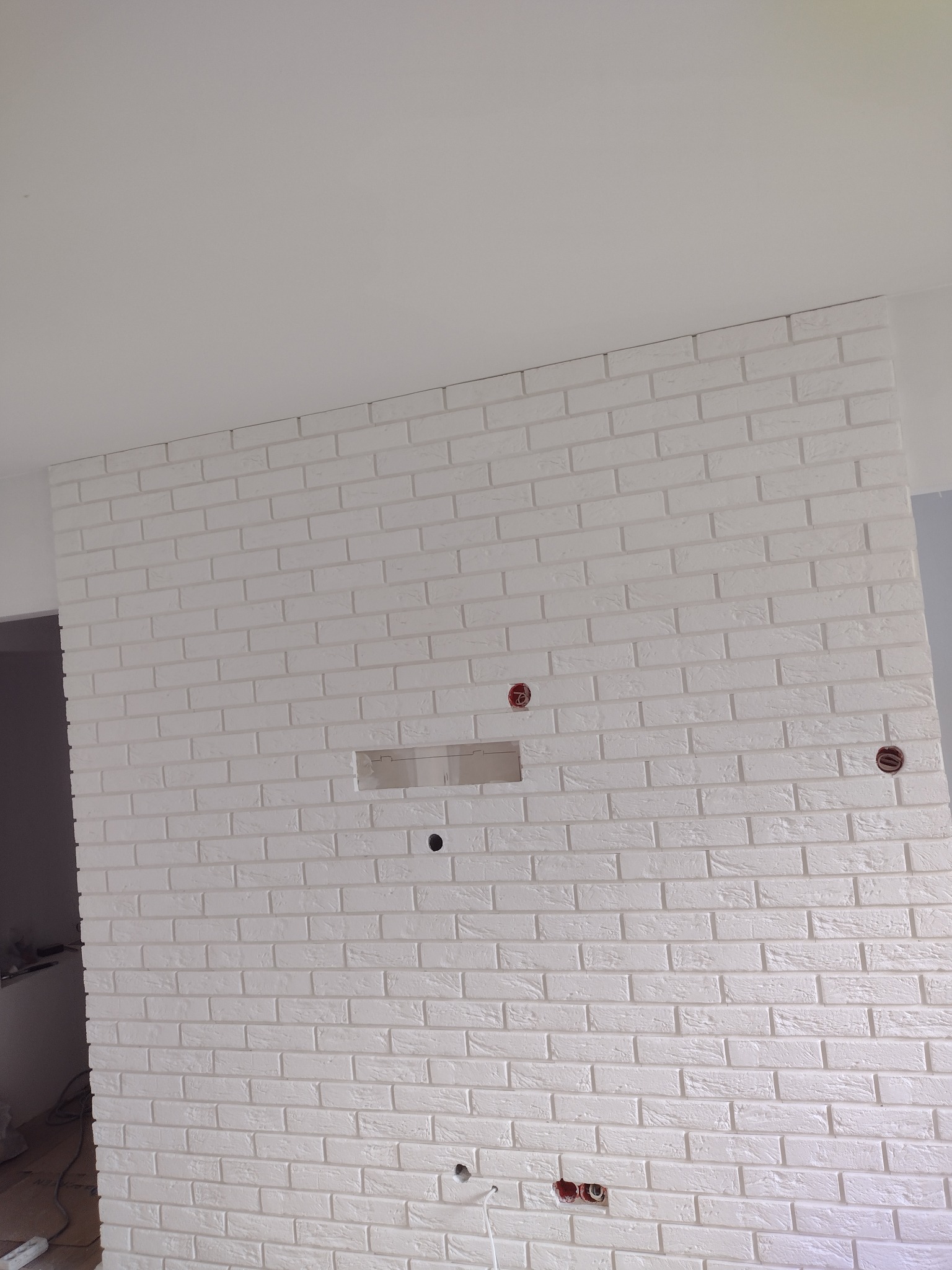 Biała ściana z cegieł dekoracyjnych z widocznymi otworami na gniazdka elektryczne i przewody, w trakcie prac wykończeniowych.