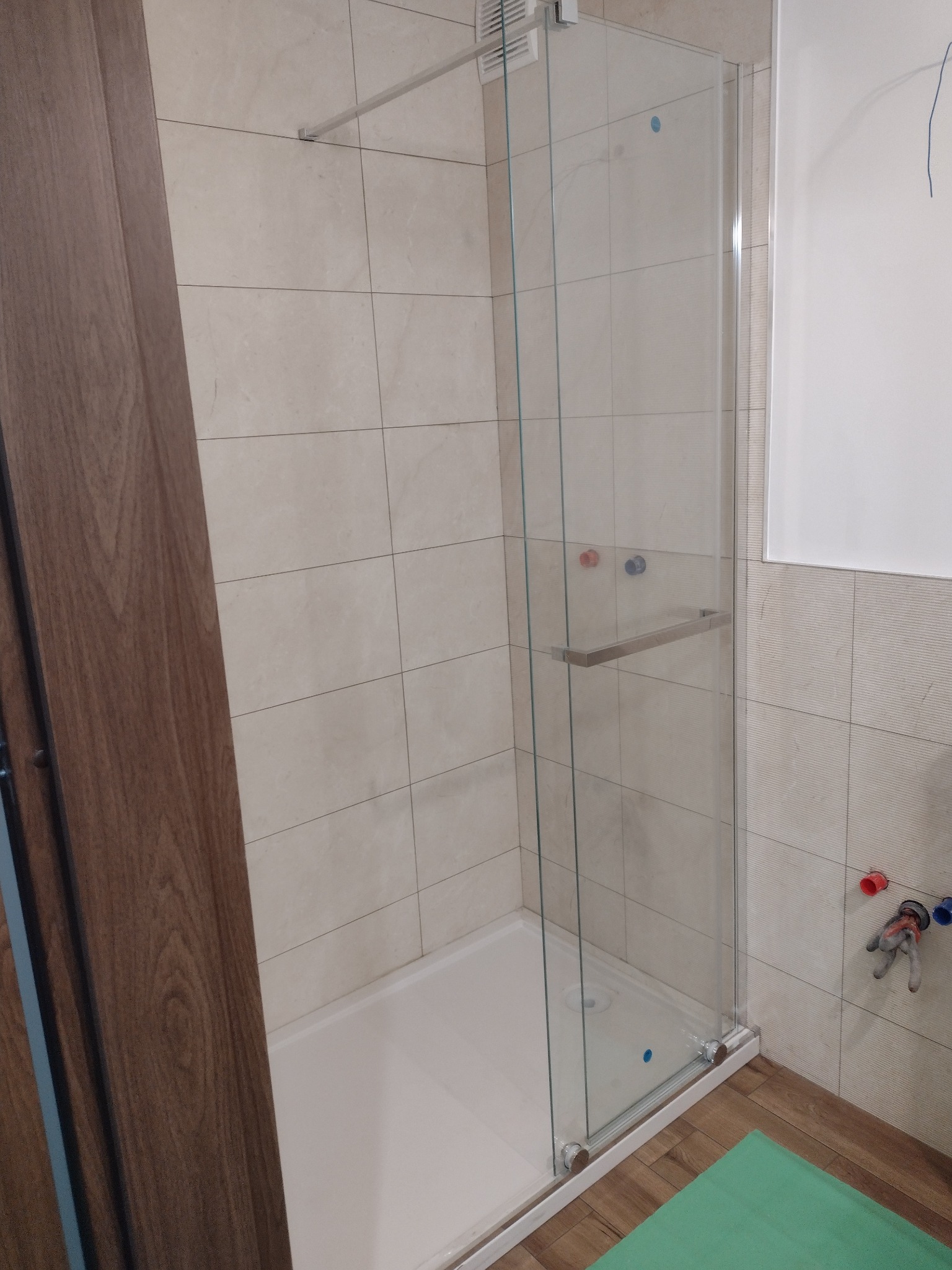 Szklana kabina prysznicowa z brodzikiem, zamontowana w łazience wyłożonej jasnymi płytkami, widoczne przyłącza wodne i fragment drewnianych drzwi.