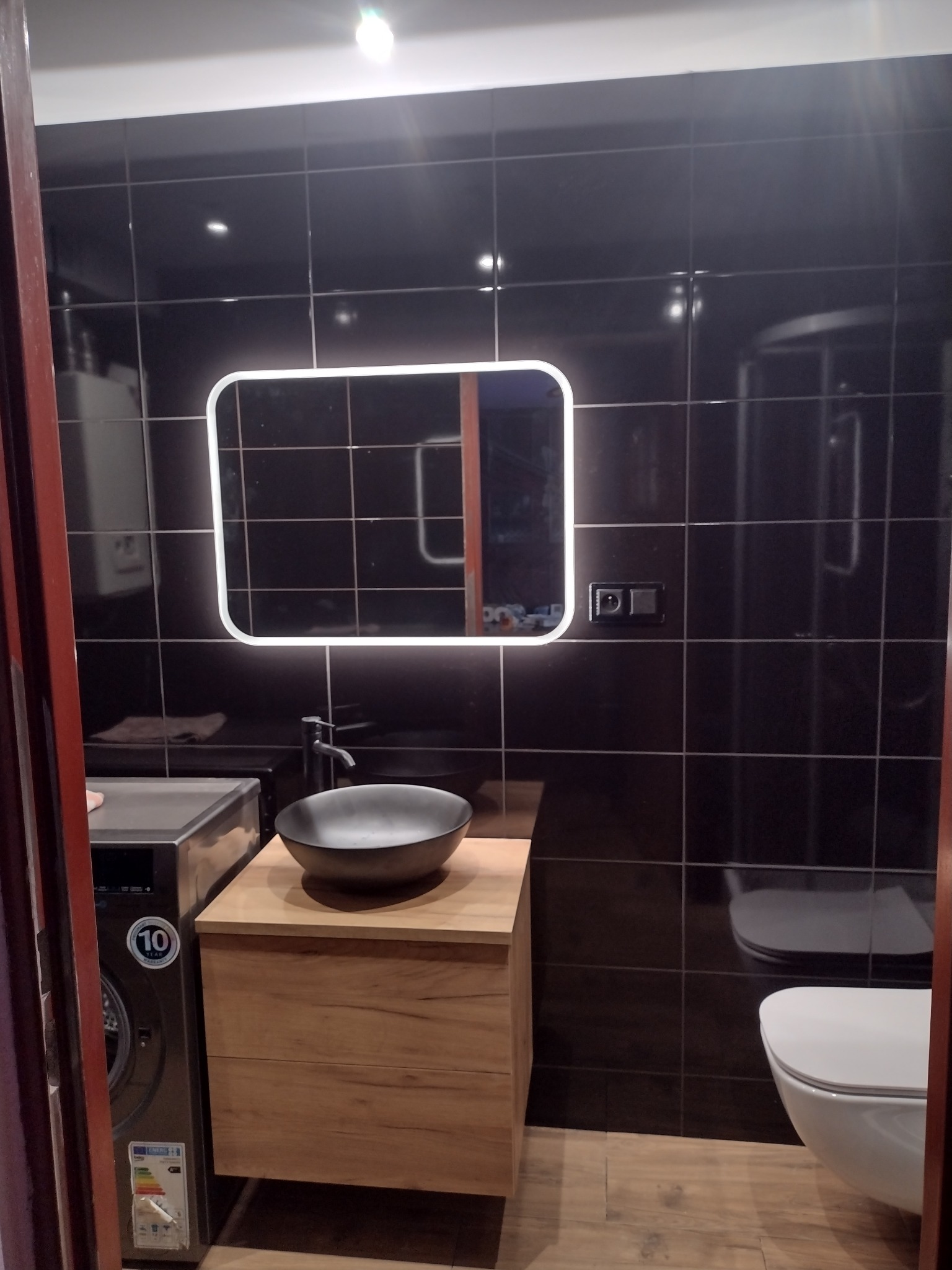 Nowoczesna łazienka z czarnymi płytkami, podświetlanym lustrem, umywalką nablatową na drewnianej szafce i pralką obok.