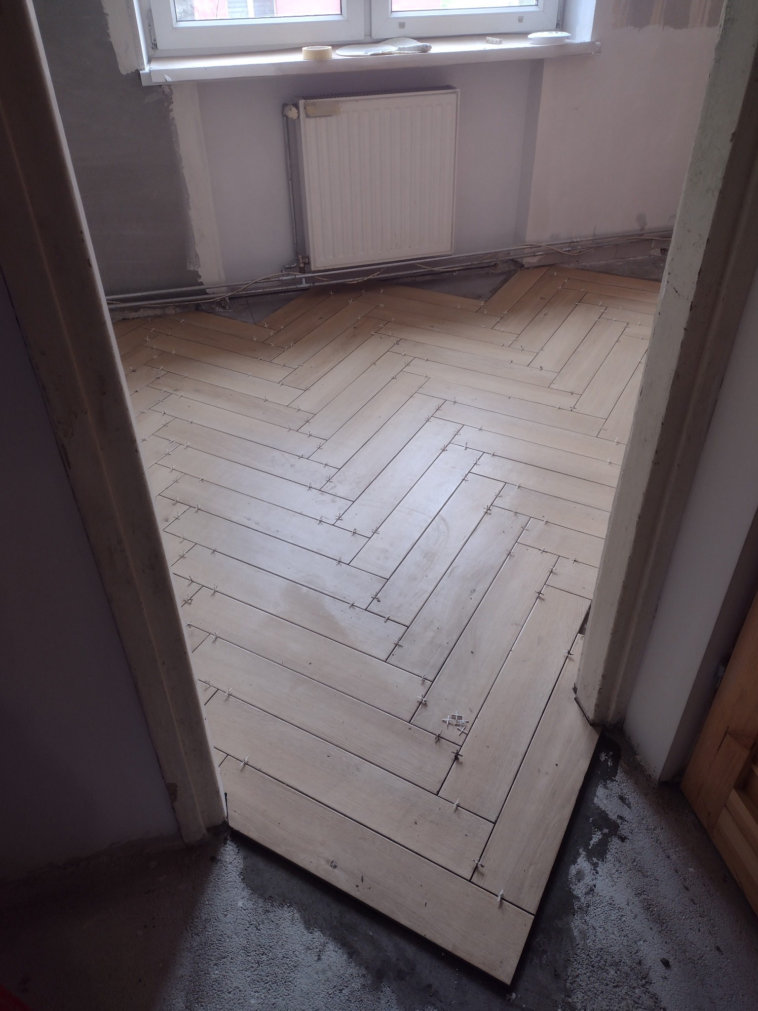 Kątowe ułożenie jasnych paneli podłogowych w trakcie montażu, widoczne krzyżyki dystansowe, surowe ściany i grzejnik w tle.