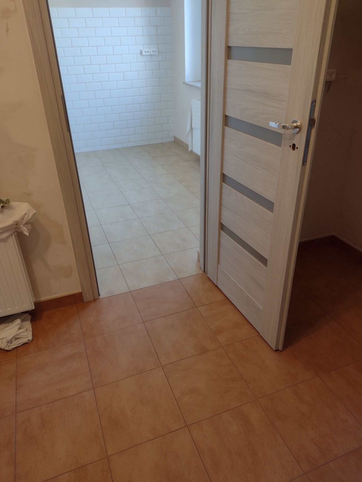 Otwarta, drewniana, wewnętrzna ościeżnica z poziomymi, szarymi wstawkami, widok na wykafelkowaną łazienkę z białymi płytkami na ścianach i beżowymi na podłodze.