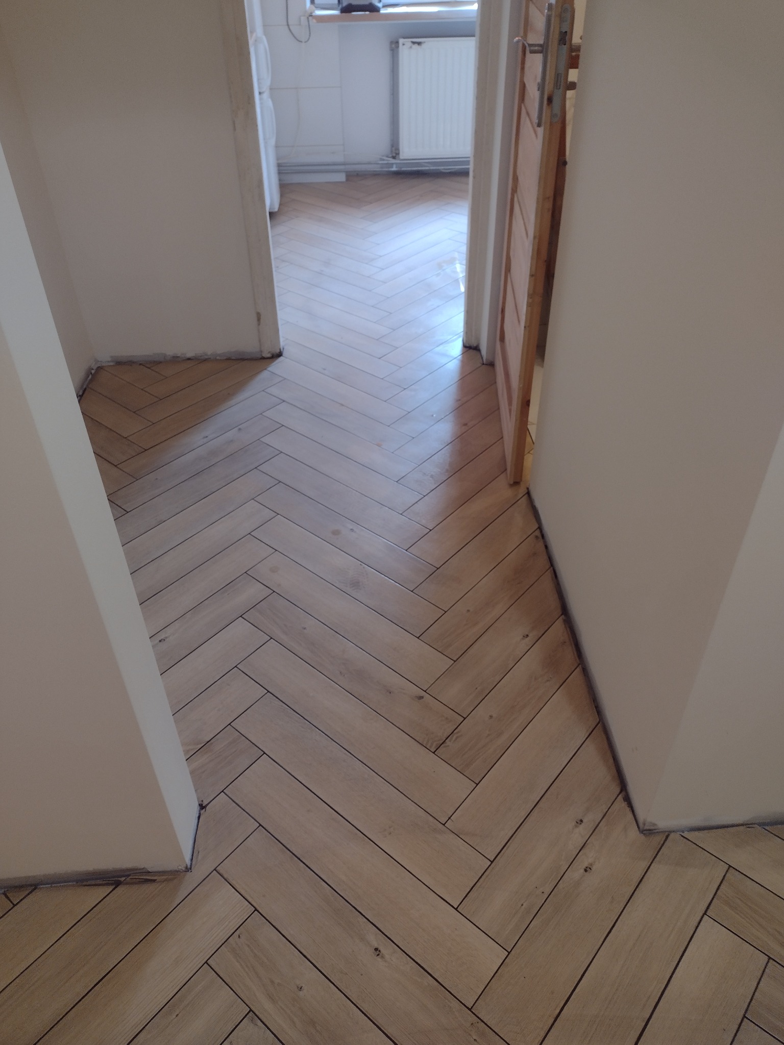 Ułożona pod kątem, jasna podłoga z paneli imitujących drewno w przedpokoju z otwartymi drzwiami i widokiem na jasną kuchnię.