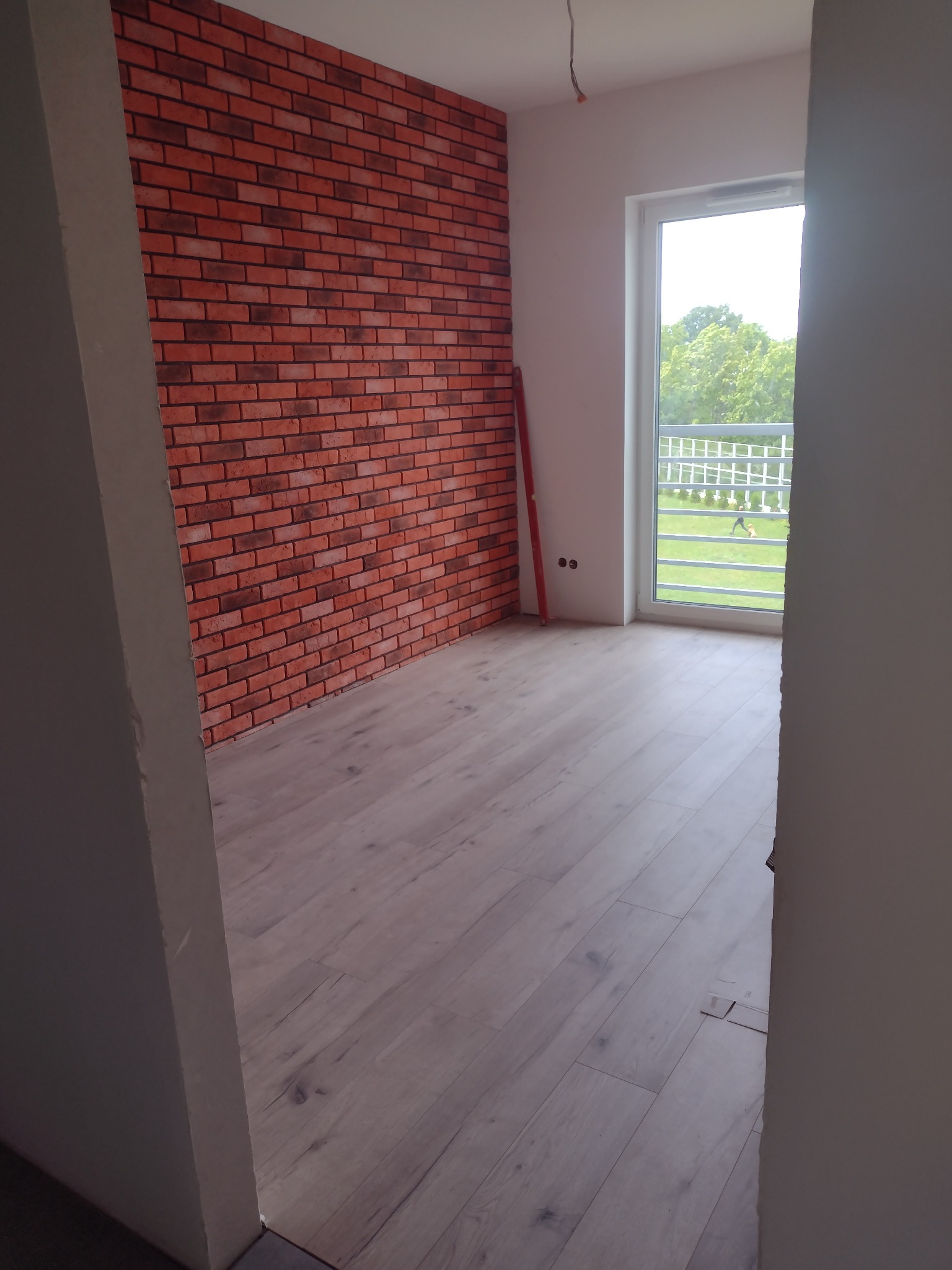 Pokój po remoncie z panelami podłogowymi w odcieniach szarości i ścianą wykończoną czerwoną cegłą dekoracyjną, widok zza futryny, na zewnątrz widoczny balkon i zieleń.