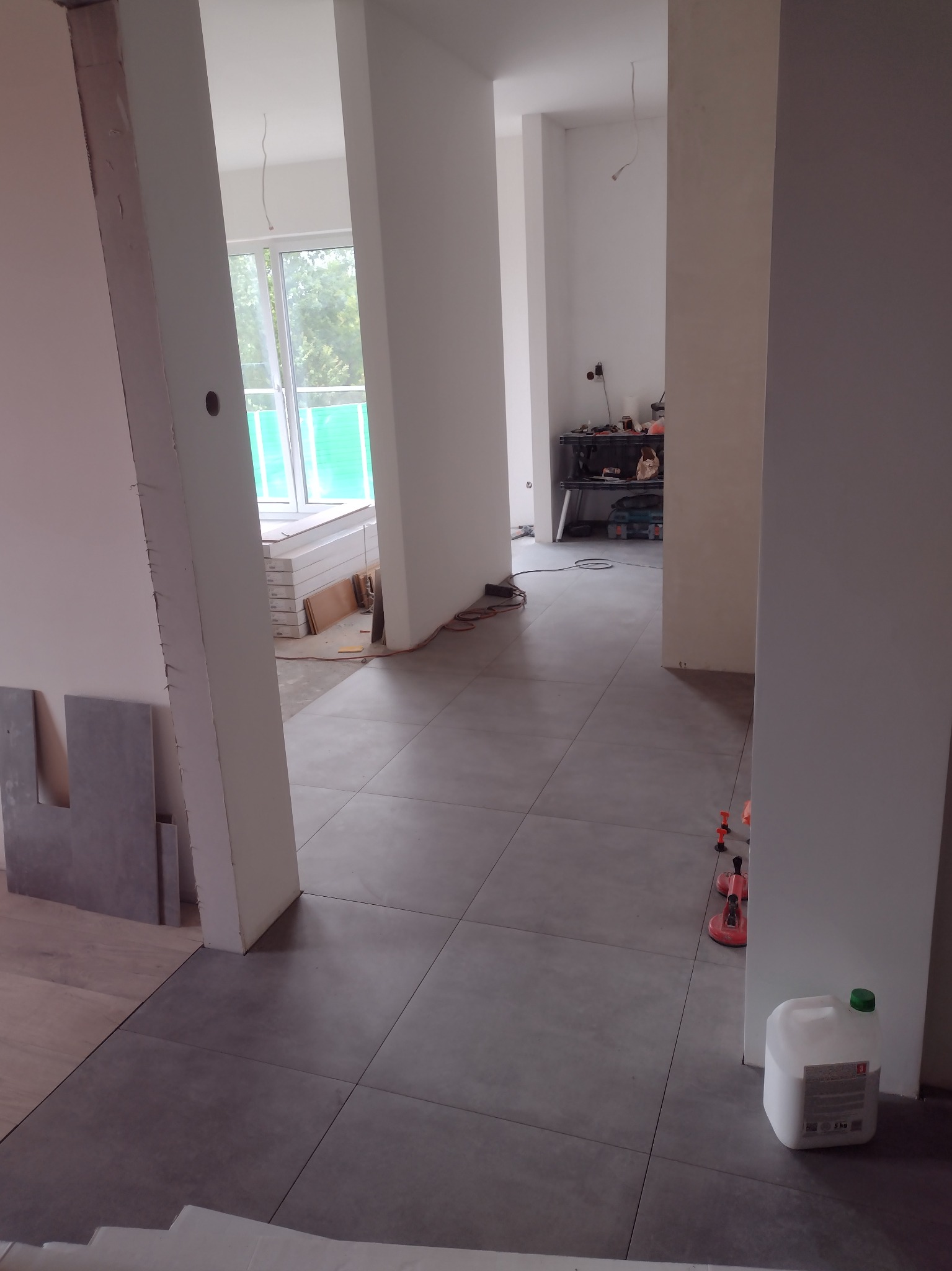 Ułożone szare płytki podłogowe w trakcie remontu, widoczne narzędzia glazurnicze, biały pojemnik z klejem i przygotowane płyty karton-gips w tle.