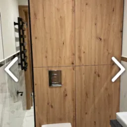 Nowoczesna łazienka z zabudową drewnopodobną kryjącą stelaż WC, wisząca miska toaletowa, marmurowe płytki podłogowe i ścienne oraz czarny grzejnik drabinkowy.