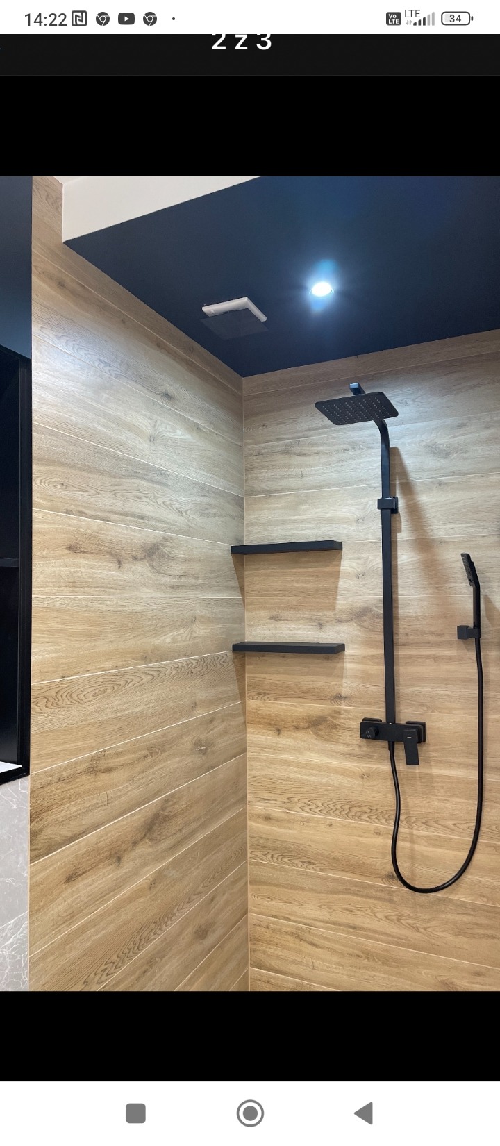 Nowoczesna kabina prysznicowa z drewnopodobnymi płytkami ułożonymi poziomo, czarną armaturą i półkami w kolorze sufitu.