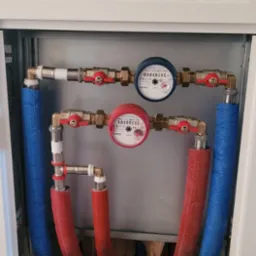 Zabudowa z dwoma wodomierzami, rury w izolacji termicznej czerwonej i niebieskiej, zawory kulowe z czerwonymi uchwytami, instalacja połączona mosiężnymi kształtkami.