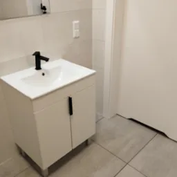 Nowoczesna łazienka z białą umywalką nablatową na szafce z czarną armaturą i uchwytami, jasne płytki na ścianach i podłodze, białe drzwi.