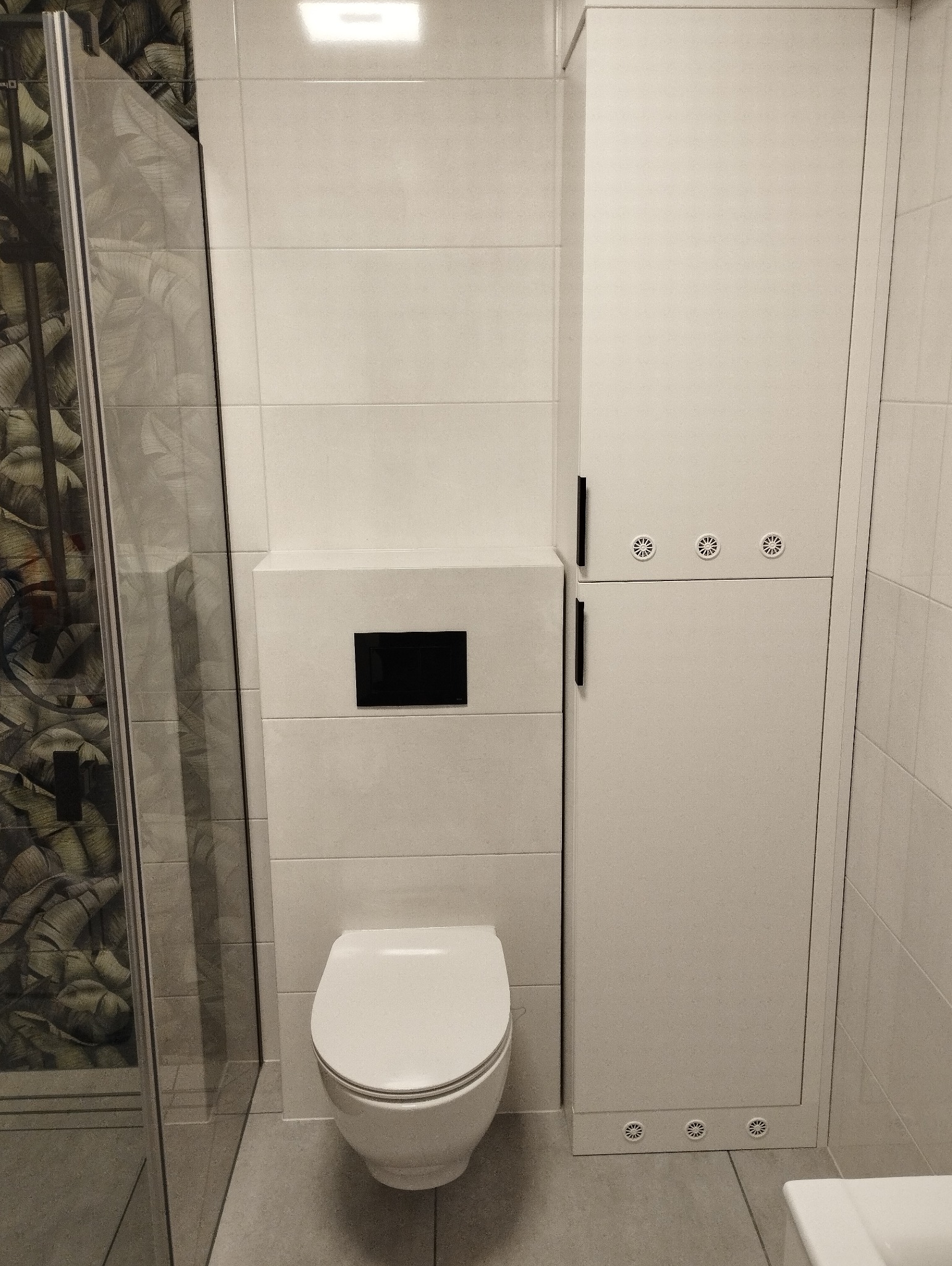 Nowoczesna, wykafelkowana łazienka z wiszącą toaletą z czarnym przyciskiem spłukiwania i wysoką, białą szafką z wentylacją u dołu, widok na kabinę prysznicową z wzorem liści.