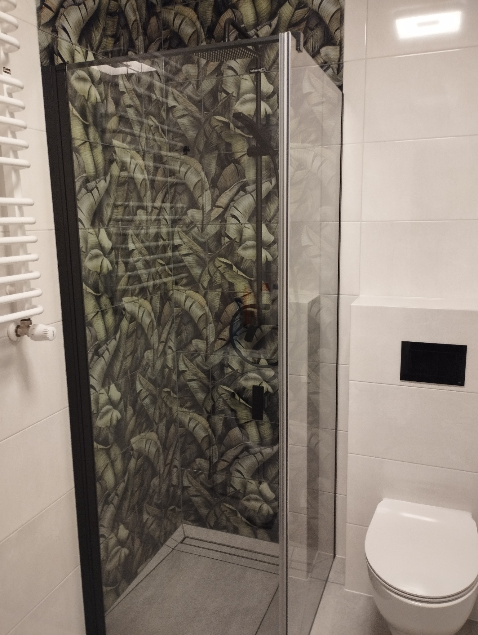Nowoczesna łazienka z kabiną prysznicową typu walk-in z czarną ramą i deszczownicą, ściana w kabinie wykończona płytkami z motywem liści bananowca, białe płytki na pozostałych ścianach, biała...