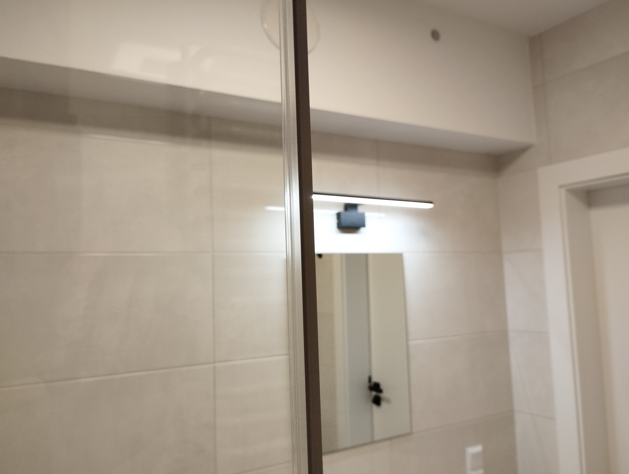 Beżowe płytki w łazience, widoczna kabina prysznicowa z brązową ramą, lustro z oświetleniem LED, oraz fragment drzwi.