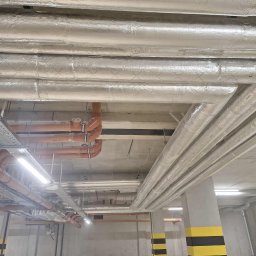 SUNOCO SPÓŁKA Z OGRANICZONĄ ODPOWIEDZIALNOŚCIĄ - Instalacja rur wentylacyjnych i hydraulicznych pod sufitem w garażu podziemnym. Rury izolowane folią aluminiową, pomarańczowe rury PVC, betonowy sufit i słupy.