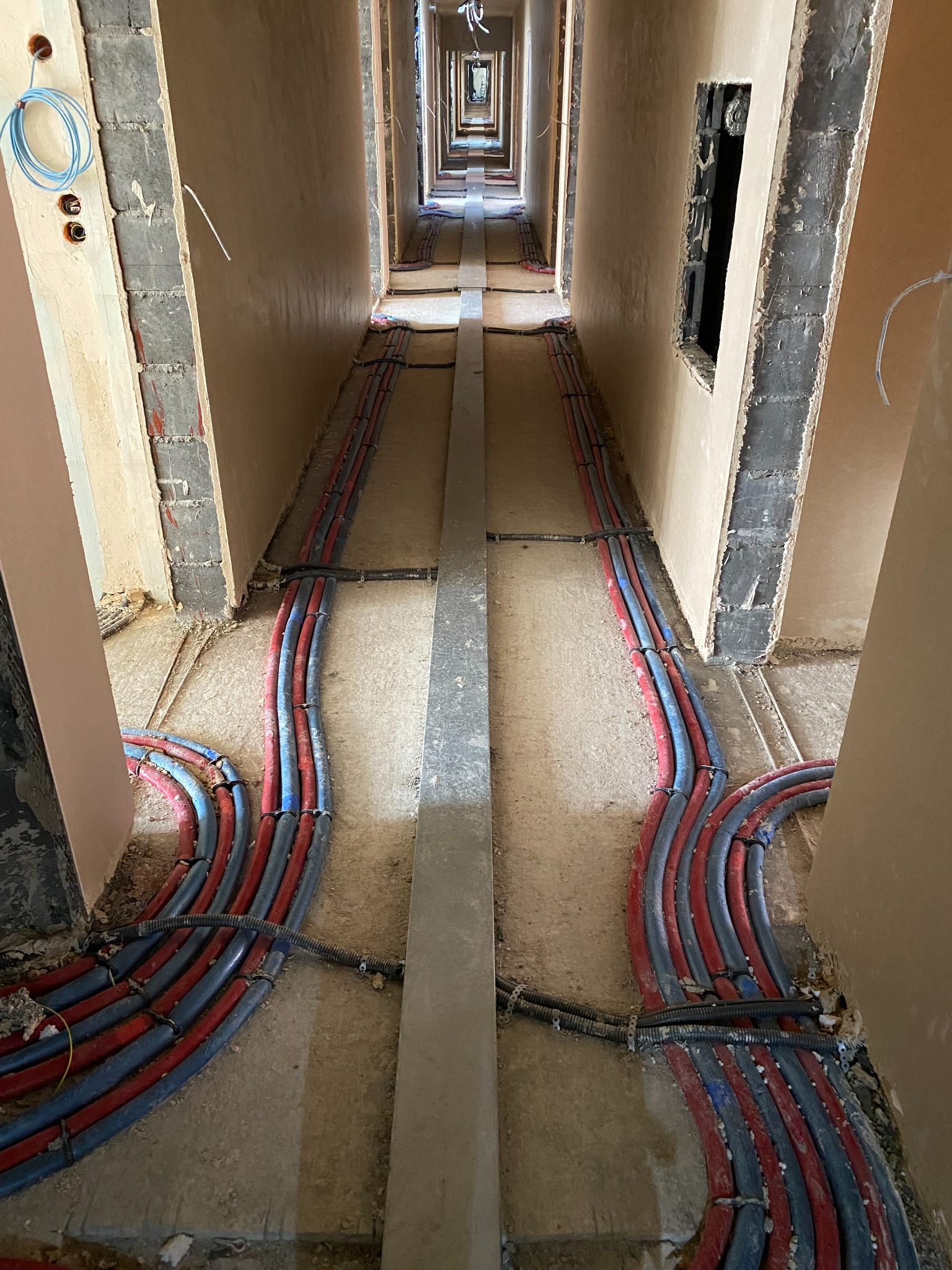 Długi korytarz w trakcie remontu z widocznymi instalacjami ogrzewania podłogowego: rurki czerwone i niebieskie ułożone wzdłuż ścian, przytwierdzone do surowej podłogi, ściany w stanie surowym...
