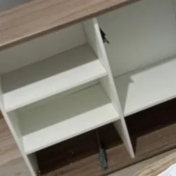 Wnętrze szafki z półkami, białe fronty i jasnobrązowy korpus, widoczne okucia i zawiasy, częściowo otwarte drzwiczki.